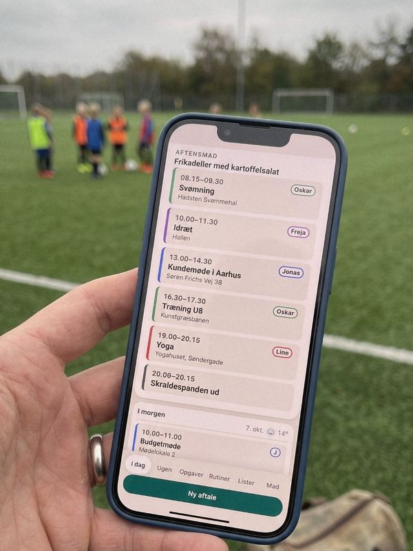 En hånd holder en telefon med familiens aftaler for i dag, blandt andet Træning U8 på kunstgræsbanen. I baggrunden står børn på en fodboldbane